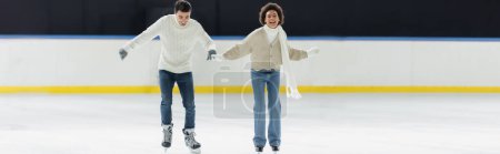 Foto de Alegre pareja interracial en guantes calientes patinaje sobre hielo en la pista, pancarta - Imagen libre de derechos