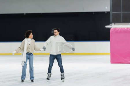 Foto de Sonriente pareja interracial tomados de la mano y mirándose mientras patina sobre hielo en la pista de patinaje - Imagen libre de derechos