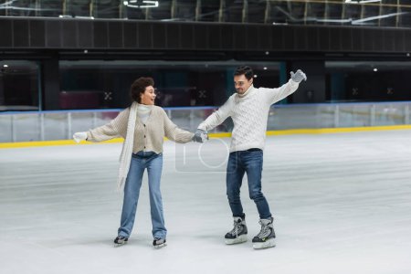 Foto de Pareja multiétnica positiva en guantes patinaje sobre hielo y divertirse en la pista de patinaje - Imagen libre de derechos