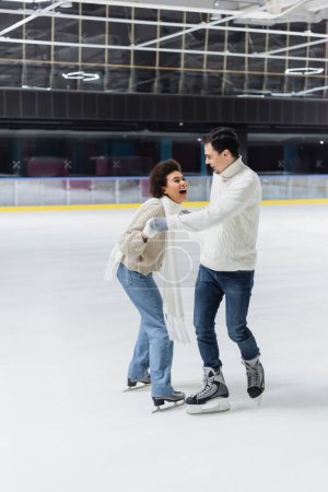 Photo pour Femme afro-américaine excitée tenant la main de son petit ami tout en patinant sur la patinoire - image libre de droit