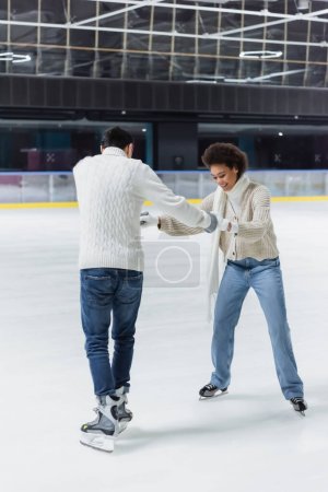 Smiling african american woman holding hands of boyfriend in ice skates on rink 