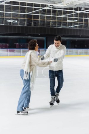 Cheerful interracial couple in jeans and sweaters holding hands while ice skating on rink 