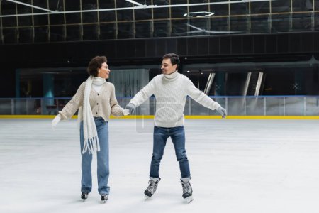 Smiling african american woman in jeans and sweater ice skating with boyfriend on rink 