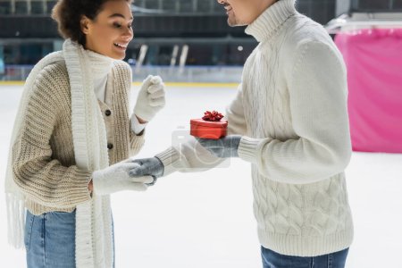 Smiling man holding heart shaped box and hand of african american girlfriend on ice rink 