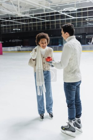 Smiling african american woman looking at heart shaped gift near boyfriend on ice rink 