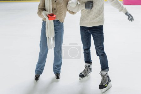 Cropped view of woman in gloves holding heart shaped gift near boyfriend on ice rink 