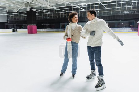 Smiling african american woman holding gift box and looking at boyfriend on ice rink 