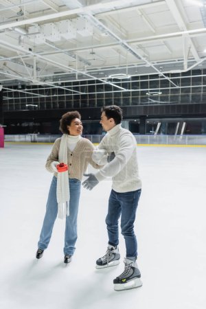 Photo for Positive african american woman holding red gift and looking at boyfriend on ice rink - Royalty Free Image