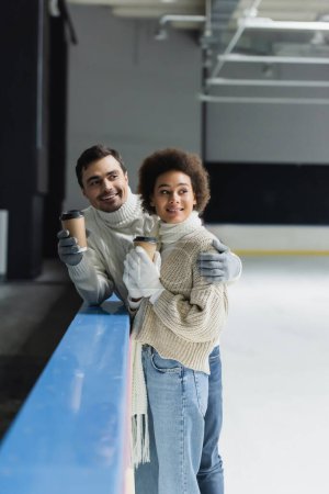 Téléchargez les photos : Homme souriant étreignant petite amie afro-américaine avec du café pour aller sur la patinoire - en image libre de droit