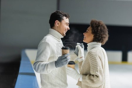 Foto de Vista lateral de la sonriente pareja interracial cogida de la mano y café para ir en pista de hielo - Imagen libre de derechos