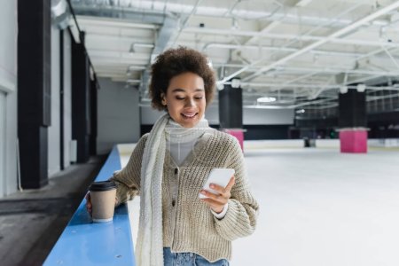 Foto de Mujer afroamericana sonriente en bufanda usando teléfono inteligente y sosteniendo café para ir en pista de hielo - Imagen libre de derechos