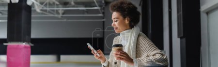 Téléchargez les photos : Vue latérale d'une femme afro-américaine tenant un café à emporter et utilisant un smartphone sur une patinoire, une bannière - en image libre de droit