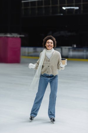 Téléchargez les photos : Femme afro-américaine positive tenant du café pour aller pendant le patinage sur glace et regardant la caméra sur la patinoire - en image libre de droit
