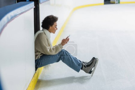 Foto de Vista lateral de mujer afroamericana en patines sobre hielo usando smartphone en pista de patinaje - Imagen libre de derechos