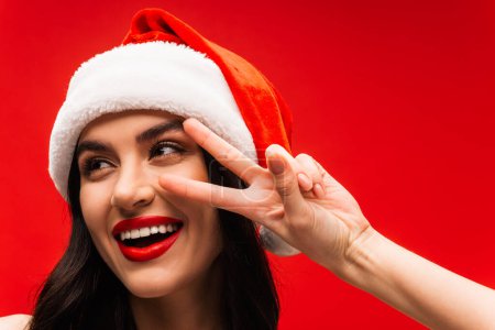 Cheerful young woman in santa hat showing victory sign isolated on red