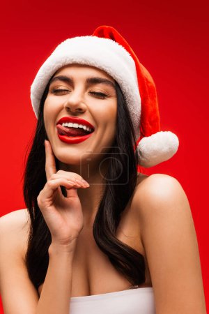 Brünette Frau im Top und Weihnachtsmannmütze, die herausstehende Zunge isoliert auf Rot