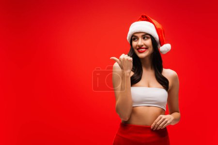 Smiling woman in santa hat pointing with finger isolated on red