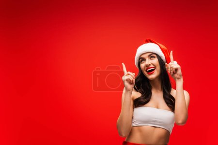 Cheerful brunette model in top and santa hat pointing with fingers isolated on red