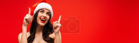 Positive brunette woman in santa hat pointing with fingers isolated on red, banner 