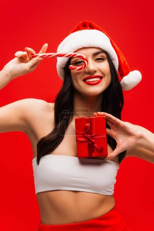 Modelo sonriente en sombrero de santa celebración de piruleta de Navidad y presente aislado en rojo