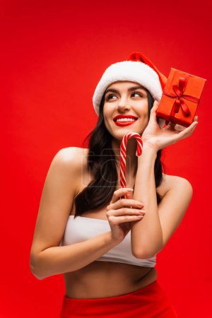 Cheerful young woman in santa hat holding striped lollipop and present isolated on red