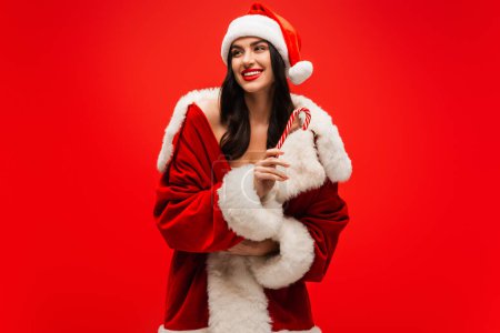 Positive brunette model in santa costume holding striped lollipop isolated on red