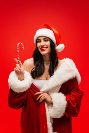 Brunette woman in santa costume looking at striped lollipop isolated on red