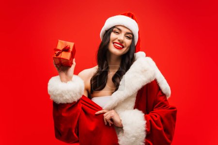 Positive model in santa costume holding present and looking at camera isolated on red