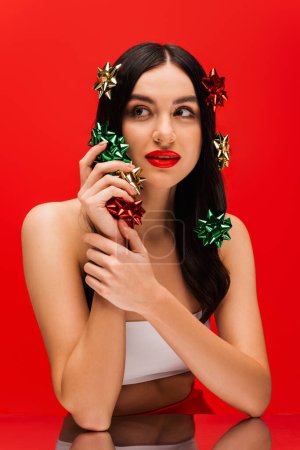 Téléchargez les photos : Jeune femme avec maquillage tenant des noeuds cadeaux près du miroir isolé sur rouge - en image libre de droit