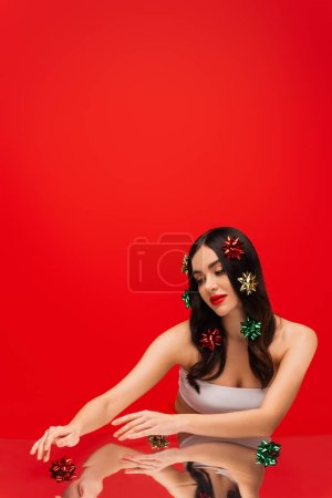 Photo for Young woman with makeup touching gift bows on reflective surface isolated on red - Royalty Free Image