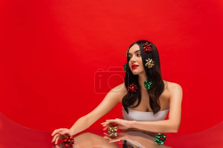 Téléchargez les photos : Jeune femme avec des épaules nues et des nœuds cadeaux sur les cheveux posant près du miroir isolé sur rouge - en image libre de droit