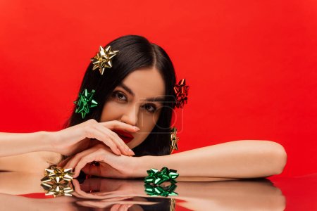 Brunette woman with gift bows on hair looking at camera near reflective surface isolated on red