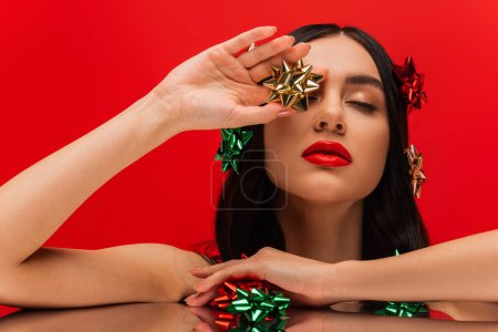 Brunette model with visage holding shiny gift bow near mirror isolated on red 
