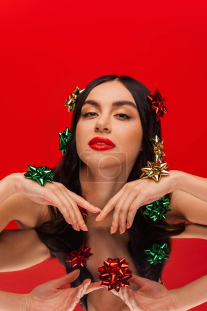 Téléchargez les photos : Modèle Brunette avec visage et noeuds cadeaux sur les cheveux posant près du miroir isolé sur rouge - en image libre de droit