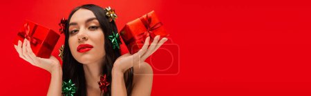 Photo for Young woman with gift bows on hair holding presents and looking at camera isolated on red, banner - Royalty Free Image