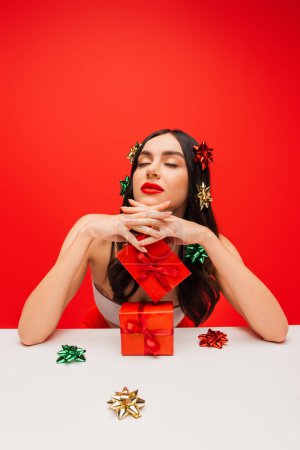 Pretty woman in top and gift bows on hair posing near presents isolated on red 