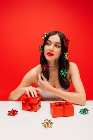Pretty young woman with gift bows on hair looking away near presents isolated on red 
