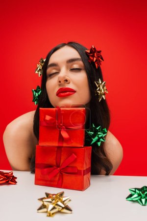 Photo for Brunette model with shiny gift bows on hair posing near presents isolated on red - Royalty Free Image