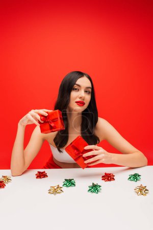 Téléchargez les photos : Joli modèle avec des cadeaux de tenue de visage près de noeuds cadeaux isolés sur rouge - en image libre de droit