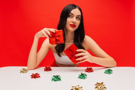 Brunette woman with makeup holding presents near shiny gift bows isolated on red 