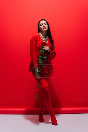 Full length of stylish young woman holding rose on red background 