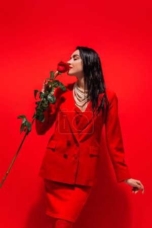 Photo for Young woman in jacket and pearl necklace smelling rose on red background - Royalty Free Image