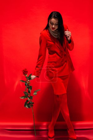 Volle Länge der stilvollen Frau in Jacke und Perlenkette mit Rose auf rotem Hintergrund 