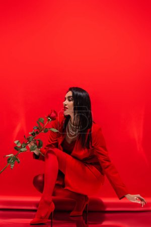 Stylish woman in heels and pearl necklace holding rose on red background 