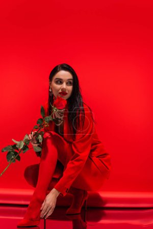 Photo for Young brunette woman with hairstyle and makeup holding rose on red background - Royalty Free Image