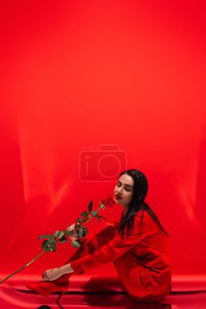 Elegante Frau in High Heels duftet Rose, während sie auf rotem Hintergrund sitzt 