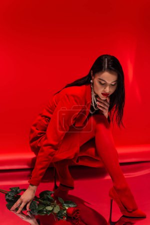 Photo for Stylish model in heels and pearl necklace looking at reflective surface near rose on red background - Royalty Free Image