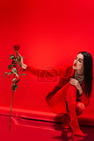 Stylish brunette woman in pearl necklace and heels looking at rose on red background 