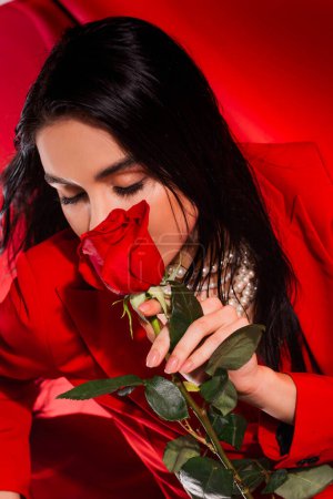 Brunette woman in pearl necklace holding rose near face on red background 