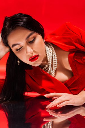 Mujer morena con estilo en collar de perlas tocando superficie reflectante sobre fondo rojo 